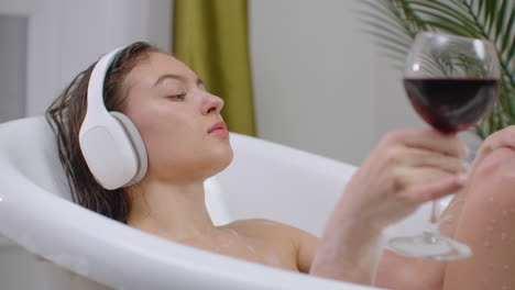 Beautiful-young-woman-enjoying-pleasant-bath-with-foam-holding-a-glass-of-wine-Woman-listening-to-music-in-bath.-Woman-listening-to-music-in-bubble-bath