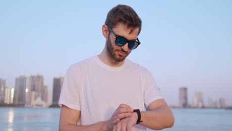 Happy-handsome-man-in-sunglasses-and-white-t-shirt-uses-a-watch-watch-looks-and-presses-his-finger-on-the-screen-standing-on-the-waterfront-in-summer-against-the-city-and-buildings