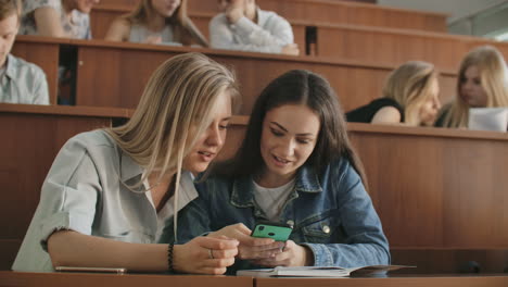 Pretty-girls-students-are-using-smartphone-watching-screen-talking-and-laughing-sitting-at-desks-at-university.-Social-media-internet-millennials-and-education-concept