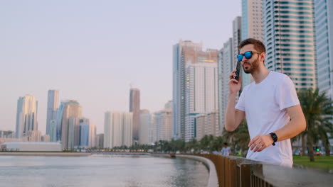 A-young-male-tourist-in-sunglasses-and-bristles-hipster-in-a-white-t-shirt-standing-on-the-waterfront-against-the-background-of-the-modern-city-writes-text-messages-phone-for-social-networks-is-live-broadcast