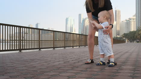 Smiling-Boy-holding-his-mother's-hand-makes-the-first-steps-walking-along-the-promenade-in-the-summer.