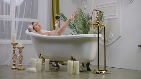 Young-brunette-woman-is-taking-foamy-bath-relaxes-and-enjoys-drinks-white-wine-girl-is-drinking-champagne-in.-Bathroom-blowing-on-foam-lady-touches-legs