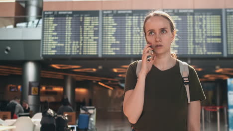 Young-beautiful-woman-standing-at-the-airport-calls-on-the-phone-on-the-background-of-the-scoreboard-with-information-about-the-departures.