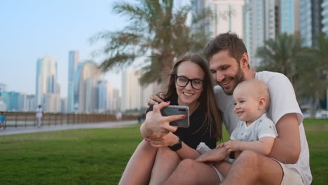 Glückliche-Familie-Mit-Zwei-Kindern,-Die-Zusammen-Auf-Dem-Gras-Im-Park-Sitzen-Und-Ein-Selfie-Mit-Smartphone-Machen.