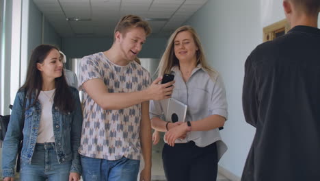 Three-students-walk-down-the-corridor-of-the-University-and-look-at-the-smartphone-screen-and-laugh.