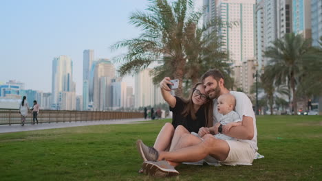 Happy-family-with-two-children-sitting-together-on-grass-in-park-and-taking-a-selfie.-With-smartphone.
