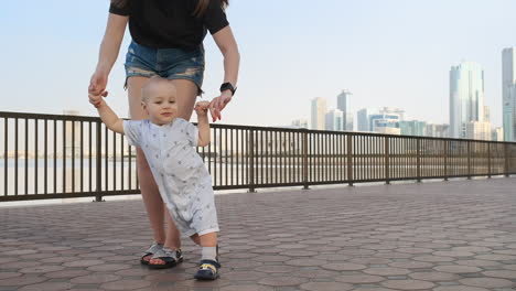 The-boy-holding-his-mother's-hand-makes-the-first-steps-walking-along-the-promenade-in-the-summer