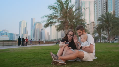 Happy-family-with-two-children-sitting-together-on-grass-in-park-and-taking-a-selfie.-With-smartphone.