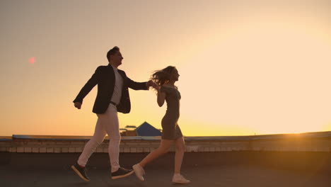 Cute-lovers-laughing-running-on-the-roof-of-the-building-at-sunset-on-the-background-of-the-city-and-hugging-standing-at-the-edge.-Romance-and-love