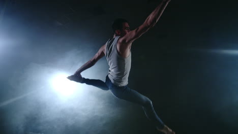 A-modern-ballet-a-man-performs-jumps-and-spins-in-the-light-of-spotlights-and-smoke-on-a-dark-background.-Acrobatic-choreography-rehearsal-of-the-script-of-modern-ballet.