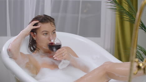 young-sexy-woman-with-straight-naked-body-and-clean-legs-lying-and-relaxing-in-white-foam-bath-tub-with-candles-around-in-light-bathroom-drink-alcohol-from-wine-glass-indoors