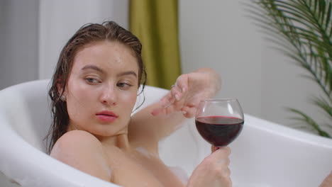 A-seductive-brunette-lies-in-a-bath-with-foam-relaxes-and-drinks-red-wine.-Recuperate-and-relax-in-a-hot-bubble-bath.