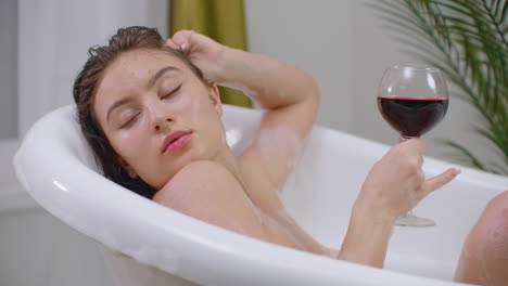 A-beautiful-young-brunette-woman-lies-in-a-bubble-bath-relaxing-and-drinking-red-wine.-Recuperate-and-relax-in-a-hot-bubble-bath.