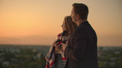 A-young-couple-on-the-roof-drinking-wine-from-glasses-standing-in-a-blanket-and-admiring-the-beautiful-sunset-over-the-city.-Romantic-evening-on-the-roof-overlooking-the-city