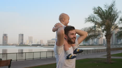Loving-Father-in-a-white-t-shirt-in-the-summer-walks-with-a-child-sitting-on-the-neck-against-the-city-and-palm-trees