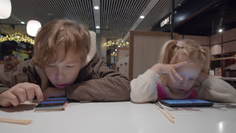 Niños-Usando-Teléfonos-Mientras-Esperan-Comida-En-La-Cafetería