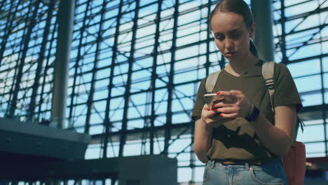 A-female-traveler-with-a-backpack-stands-at-the-airport-looking-at-the-smartphone-screen-and-typing-a-message.-Electronic-boarding-pass-for-plane-train
