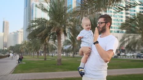 Father-playing-with-sons-in-the-summer-in-a-modern-city-holding-him-and-hugging-standing-in-a-white-t-shirt-and-shorts