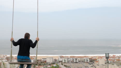 Man-swinging-on-the-hill-overlooking-resort-town-and-ocean-Nazare-in-Portugal