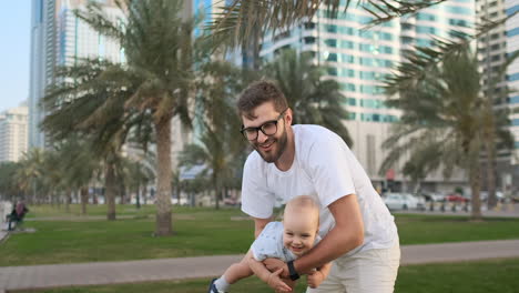 Father-playing-with-sons-in-the-summer-in-a-modern-city-holding-him-and-hugging-standing-in-a-white-t-shirt-and-shorts