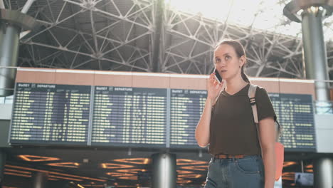 Standing-at-the-display-with-information-about-the-departure-of-aircraft-at-the-airport-a-young-girl-with-a-backpack-talking-on-a-mobile-phone