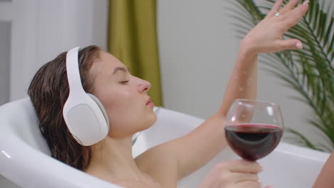 A-young-beautiful-Caucasian-brunette-lies-in-the-bathroom-and-listens-to-music-in-white-headphones-resting-from-stress-and-relaxing-drinking-red-wine-from-glass