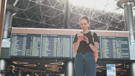 Young-beautiful-woman-traveler-standing-at-the-airport-with-a-backpack-and-looking-at-the-smartphone-screen