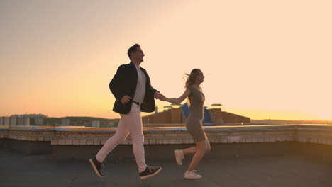 Young-couple-holding-hands-walking-woman-leading-boyfriend-the-roof-of-the-building-at-sunset-POV-travel-concept.-Carefree-free-lovers-run-on-the-roof-laughing-and-smiling