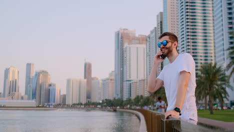 Young-male-tourist-in-sunglasses-and-bristles-hipster-in-white-t-shirt-standing-on-the-waterfront-in-the-background-of-modern-city-talking-on-the-phone-and-listening-to-the-answer