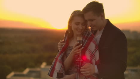 A-woman-with-glasses-of-wine-is-on-the-roof-and-a-man-puts-a-blanket-on-his-shoulders-and-hugs.-Loving-couple-drinks-wine-and-cuddles-at-sunset-on-the-roof