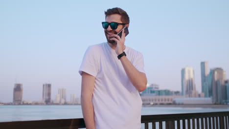 Happy-handsome-man-in-sunglasses-and-white-t-shirt-with-bristles-talking-on-the-phone-standing-on-the-waterfront-in-the-summer-against-the-city-and-buildings