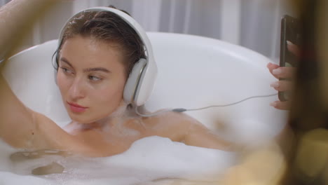 Listening-to-music-while-lying-in-a-bubble-bath-and-looking-at-the-screen-of-your-smartphone.-A-young-brunette-is-relaxing-in-the-bathroom-and-using-the-phone