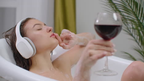 Pretty-young-woman-listening-to-music-in-headphones-while-taking-bath-and-drink-red-wine-from-a-glass.-Attractive-girl-relaxing-has-rest-at-home.-Bathroom-eco-friendly-interior.-Stress-relief-and-feel-good.-Nice-content-enjoyment