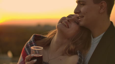 Man-and-young-pretty-woman-sitting-on-couch-holding-wine-glasses-and-kissing-on-rooftop-terrace-at-sunset