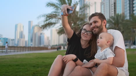 Glückliche-Familie-Mit-Zwei-Kindern,-Die-Zusammen-Auf-Dem-Gras-Im-Park-Sitzen-Und-Ein-Selfie-Mit-Smartphone-Machen.