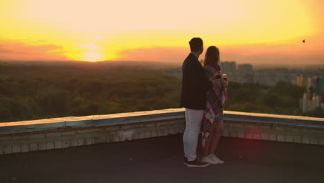 A-young-couple-on-the-roof-drinking-wine-from-glasses-standing-in-a-blanket-and-admiring-the-beautiful-sunset-over-the-city.-Romantic-evening-on-the-roof-overlooking-the-city