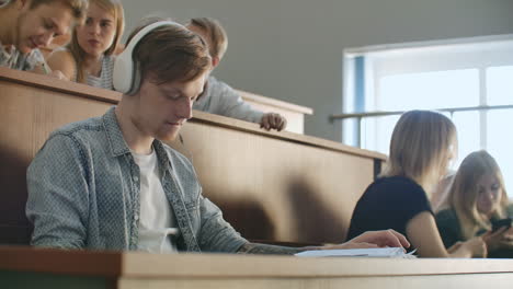 A-male-student-in-a-University-auditorium-listens-to-music-in-white-headphones-during-a-break-without-paying-attention-to-others.-A-lot-of-people-in-the-audience-are-talking.