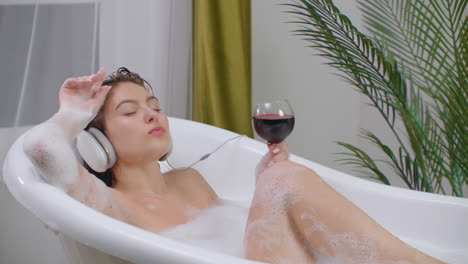 Sexy-Caucasian-brunette-in-headphones-lies-in-a-bubble-bath-and-drinks-wine-from-a-glass-and-dances-listening-to-music-in-headphones.