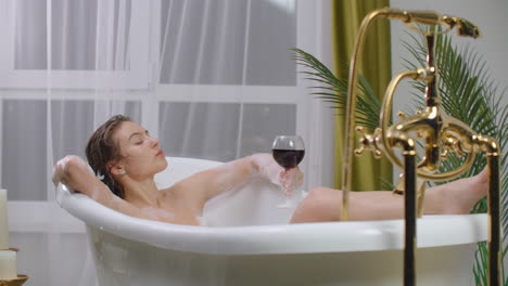 A-seductive-brunette-lies-in-a-bath-with-foam-relaxes-and-drinks-red-wine.-Recuperate-and-relax-in-a-hot-bubble-bath.