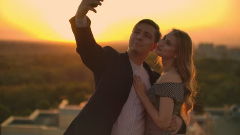 Standing-on-the-roof-at-sunset-a-married-couple-a-man-and-a-woman-hug-and-take-a-selfie-on-the-phone.-Take-pictures-of-yourself-standing-on-the-roof-and-hug.-Young-people-in-love