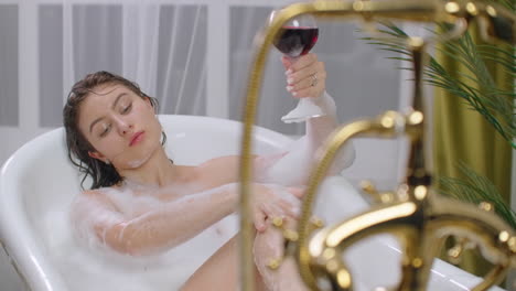 beautiful-young-brunette-woman-enjoying-pleasant-bath-with-foam-lying-with-closed-eyes-and-holding-a-glass-of-wine