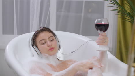 Sexy-brunette-woman-Listening-to-music-in-bath-and-drinking-wine-relaxing-and-relaxing-in-lying-bubble-bath.-Take-a-bath-after-a-hard-week.-Recuperate.