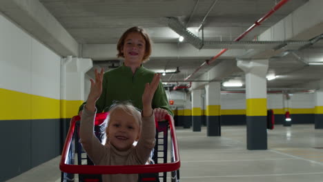 Siblings-having-fun-brother-pushing-sister-in-shopping-cart-on-supermarket-parking-lot