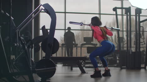The-female-fitness-instructor-in-pink-top-and-blue-leggins-is-preparing-herself-for-important-competitions.-She-is-doing-squats-with-an-empty-bar-with-a-right-technique.