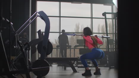 The-young-girl-in-pink-top-and-blue-leggins-is-doing-squats-with-an-empty-bar.-She-is-holding-the-bar-behind-her-neck-on-shoulders.