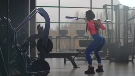 The-young-woman-in-pink-top-and-blue-leggins-is-doing-squats-with-an-empty-bar.-She-is-holding-the-bar-behind-her-neck-on-shoulders.