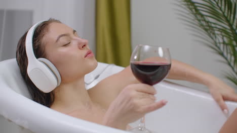 Relaxed-young-woman-listening-music-in-a-bubble-ba.-Close-up-of-a-relaxed-young-woman-listening-music-with-headphones-in-a-bubble-bath-and-drink-red-wine-from-a-glass