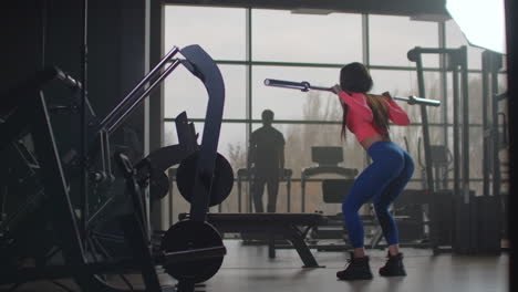 The-sports-girl-in-pink-top-and-blue-leggins-is-preparing-herself-for-important-competitions.-She-is-doing-squats-with-an-empty-bar-with-a-right-technique.