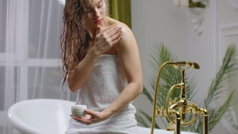 Woman-applying-cream-on-her-skin-in-bathroom-while-siting-on-bath-tub