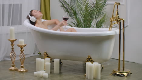 Close-up-of-young-woman-lying-in-a-bathtub-listening-to-music-with-big-headphones-on-her-head-and-drink-red-wine-from-a-glass.-Concept-of-relaxation-and-freedom.